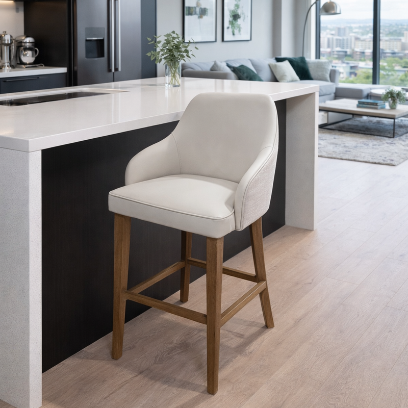A modern kitchen features a white countertop island, the Como Counter Stool with solid wood legs and light upholstery, sleek black appliances, and a bright living area framed by large windows in the background.