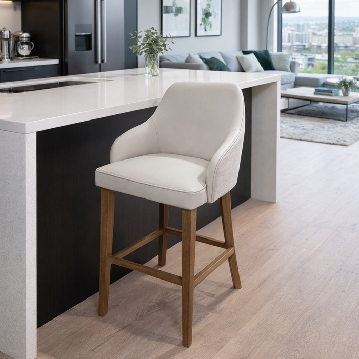 A modern kitchen features a white countertop island, the Como Counter Stool with solid wood legs and light upholstery, sleek black appliances, and a bright living area framed by large windows in the background.