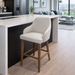 A modern kitchen features a white countertop island, the Como Counter Stool with solid wood legs and light upholstery, sleek black appliances, and a bright living area framed by large windows in the background.
