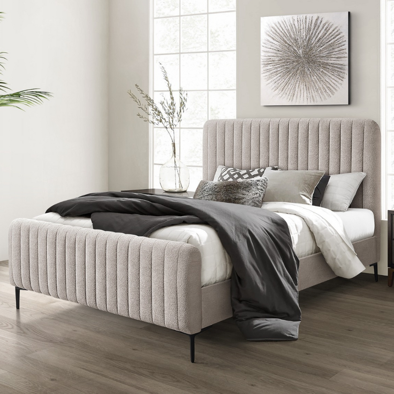 A modern bedroom featuring the Clarissa Bedframe in grey fabric with a vertically lined headboard, white and gray bedding, decorative pillows, a cozy blanket, large window, minimalist artwork, and a vase with branches.