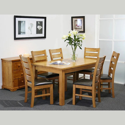 The Columbia Dining Set - Oak, with six padded chairs, surrounds a vase of white lilies and an open book. A candle-topped sideboard stands nearby, while framed art adorns the walls of this carpeted room.