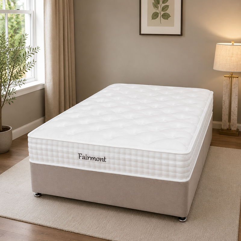 A cozy, well-lit bedroom features a neatly made bed with the Fairmont Mattress - Best Seller, known for pressure relief, set against beige walls with framed leaf artwork, a lamp, window curtains, a potted plant, and a light-colored rug.