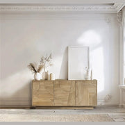 A bright room with ornate white walls, a large arched window, and the Dakota 3 Door Sideboard – Oak topped with neutral vases, dried grasses, and a blank frame.