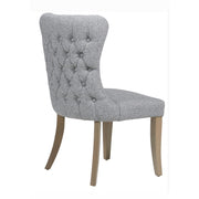 The Duke Chair is a grey upholstered dining chair featuring a button-tufted backrest and light oak legs, displayed at an angle from the back and side.