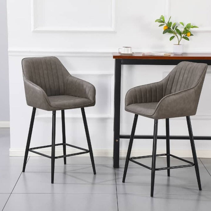 Two Enzo Counter Stools with cushioned gray seats and black metal legs are positioned in front of a tall wooden table topped with a plant, cup, and book, set against a white paneled wall.