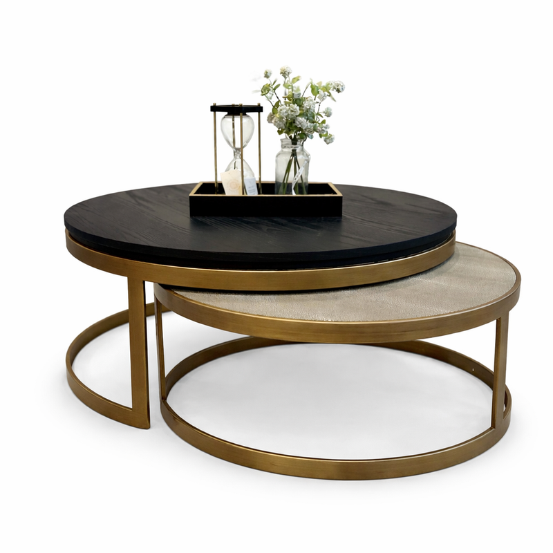 The Noir Coffee Table Nest includes two modern round tables with gold frames—a dark wood top with a tray, hourglass, and vase on the upper table, and a beige surface on the lower table.