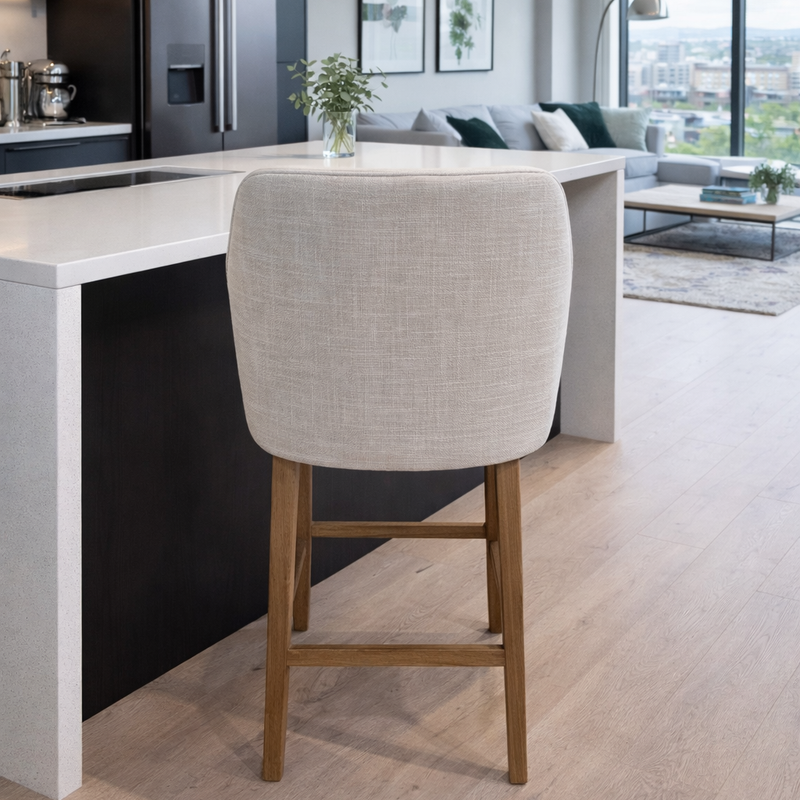 A modern kitchen features a black and white island, the Como Counter Stool with light gray upholstery and solid wood legs, and a bright living area with large windows showcasing city views in the background.