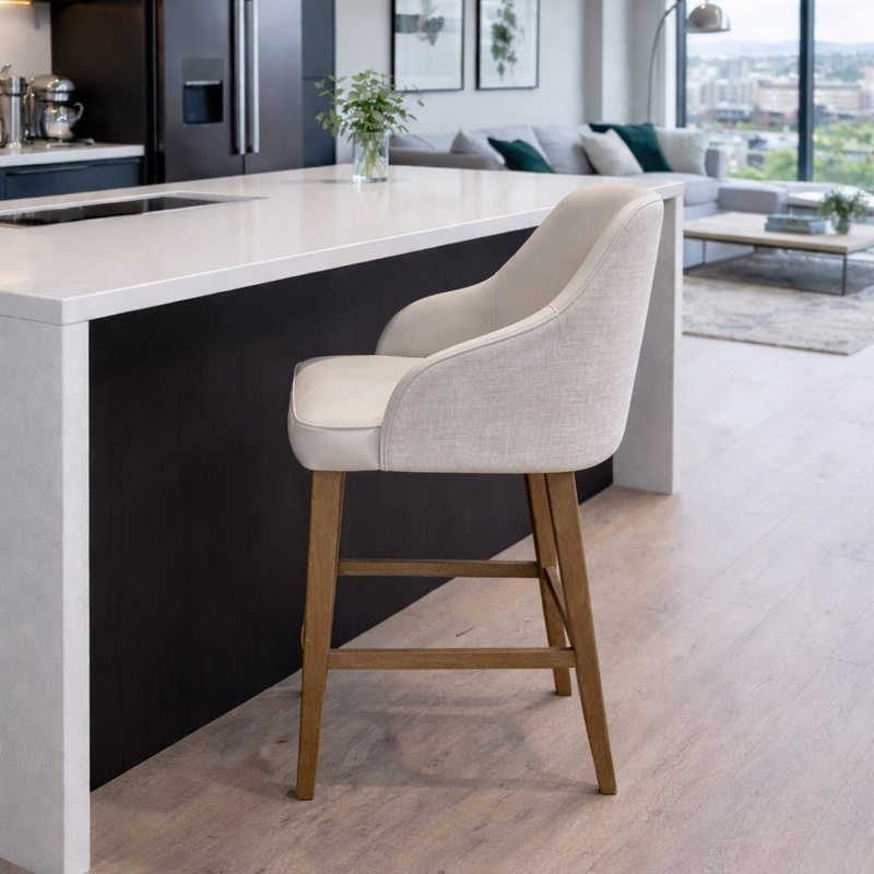 A modern kitchen with a white island, Como Counter Stools featuring wooden legs, dark cabinets, and a bright living area with large windows showcasing city views in the background.