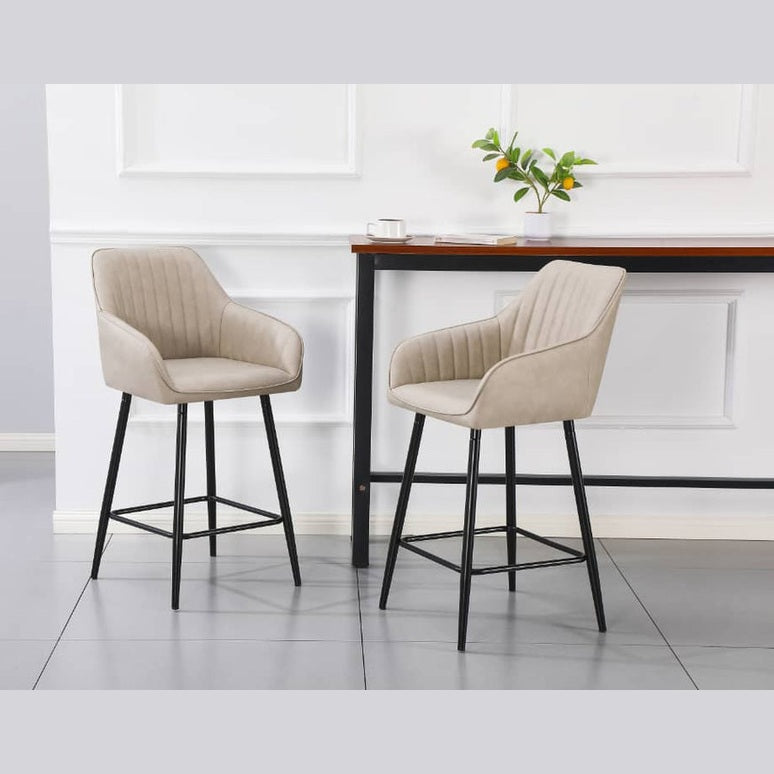 Two Enzo Counter Stools with beige upholstery and black metal legs are positioned in front of a tall wooden table against a white wall, with a small plant, cup, and book on the table for a modern minimalist look.