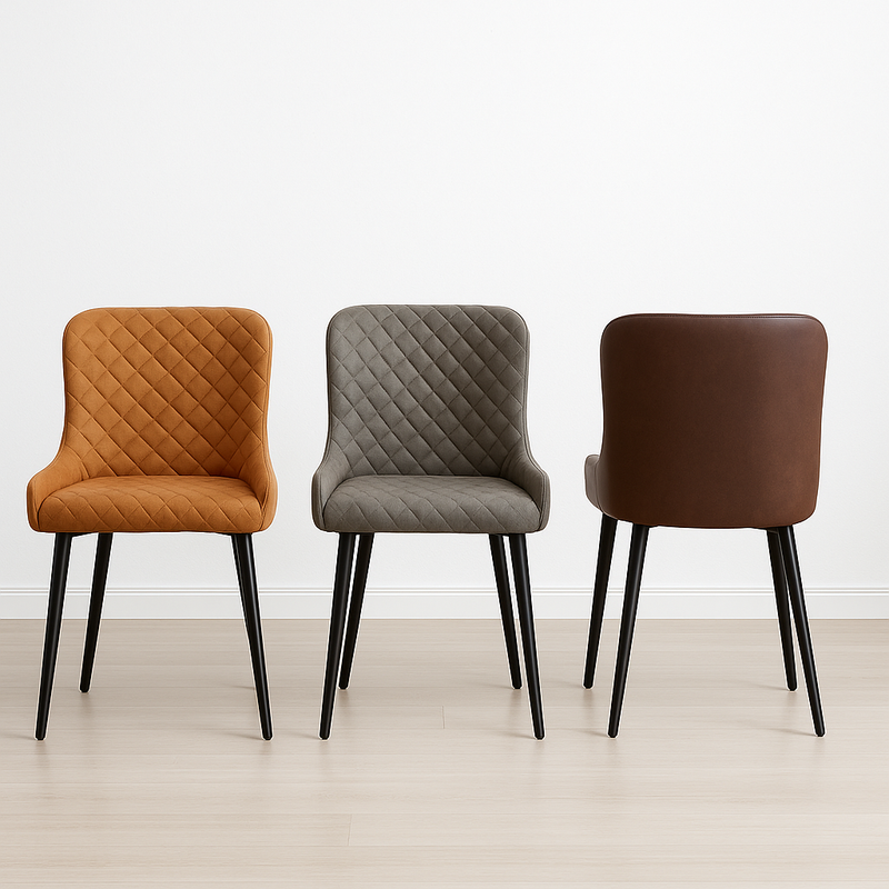 Three Diamond Dining Chairs with quilted upholstery in orange, grey PU, and brown are arranged side by side on a light wood floor before a plain white wall. The brown chair is displayed with its back facing the viewer.