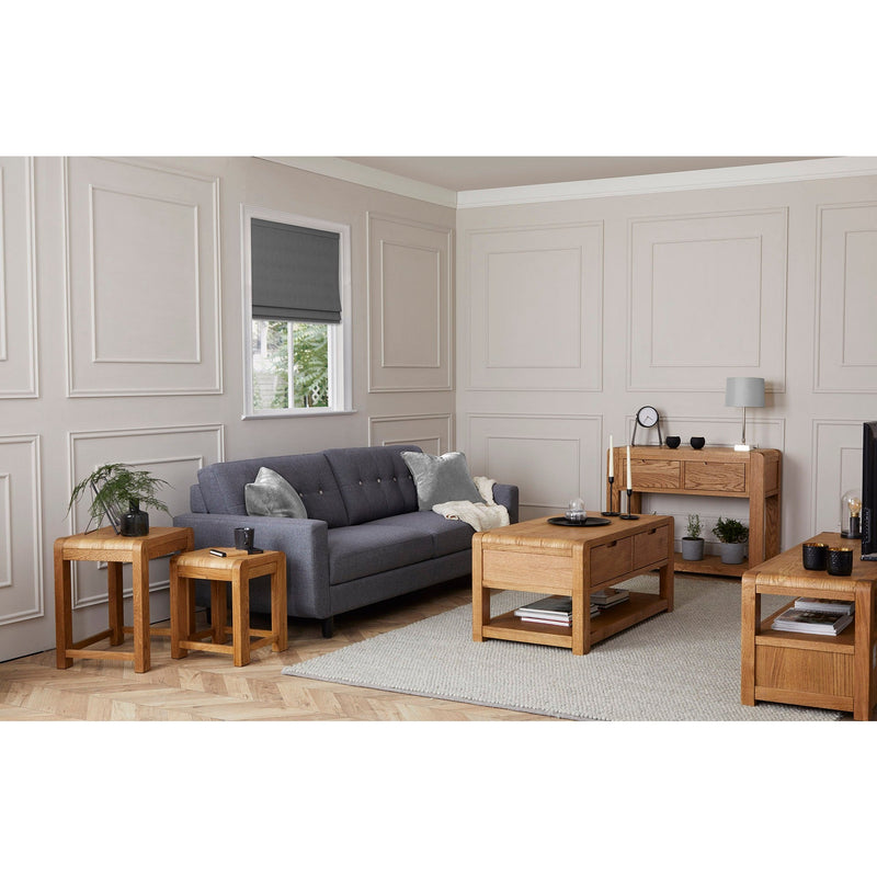 A modern living room features a gray sofa, light paneled walls, a window with a gray blind, and the Edson Nest Of Tables in solid oak on a beige rug, complemented by minimalist decor, cushions, lamps, and an oak sideboard and coffee table.