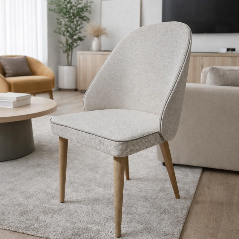 The Slim dining chair with wooden legs is placed on a light rug in a stylish living room featuring neutral decor, a coffee table, sofa, wall-mounted TV, and a potted plant by the window.