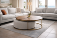 A modern living room with light gray sofas, the Todi Round Coffee Table & Stool Set featuring a gold frame and wooden top, a beige rug, and large windows revealing palm trees and ocean views.