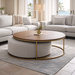 A modern living room with light gray sofas, the Todi Round Coffee Table & Stool Set featuring a gold frame and wooden top, a beige rug, and large windows revealing palm trees and ocean views.