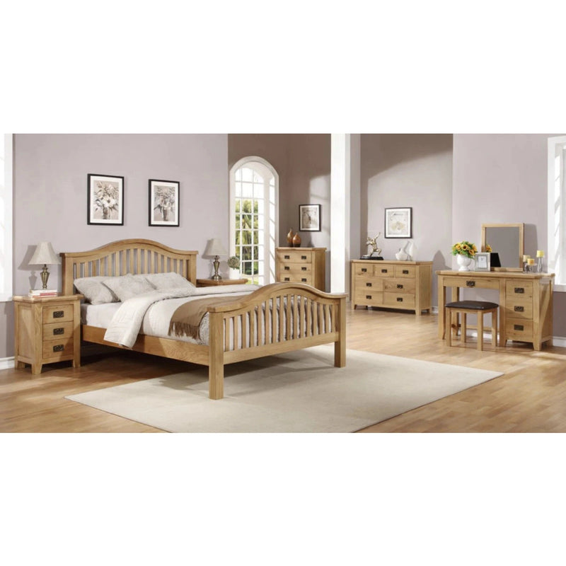 A spacious bedroom featuring the timeless Saoirse Bedframe with a curved headboard, complemented by nightstands, dressers, a vanity, pale walls, framed art, lamps, a beige rug, and large windows for an inviting atmosphere.