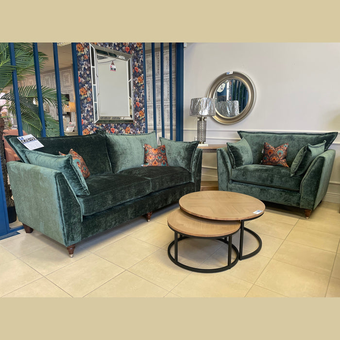 A green velvet Cleo Range sofa and armchair with deep seats and patterned cushions are paired with round wooden coffee tables, a mirror, table lamp, and decorative wall accents for an elegant showroom style.