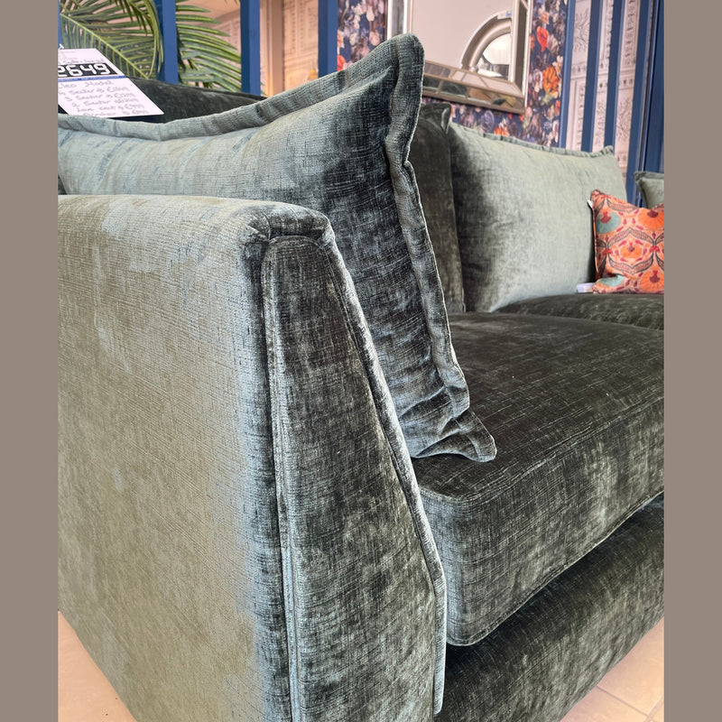 Close-up of a dark green velvet sofa from the Cleo Range, featuring deep seats and plush cushions. The stylish room has floral wallpaper, blue paneling, a mirror, a patterned throw pillow, and a price tag on the sofa's top corner.