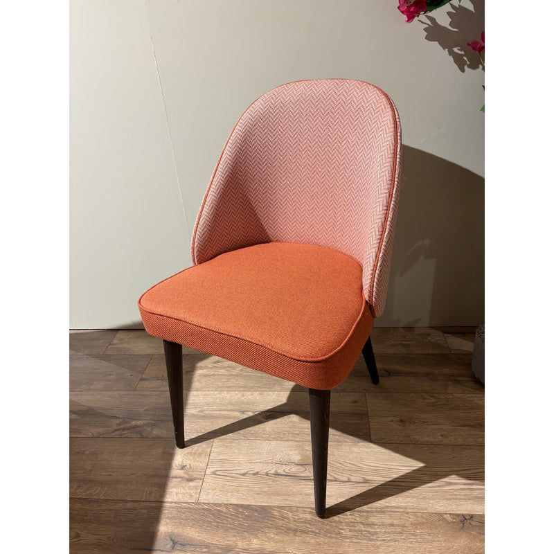 The Slim dining chair features an orange seat cushion, a light pink patterned backrest, and dark wooden legs. It sits on a wooden floor near a light wall, with pink flowers partly visible in the upper right corner.