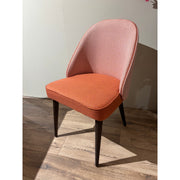 The Slim dining chair features a textured pinkish backrest and a solid orange upholstered seat cushion, supported by four dark wooden legs, and is shown on a sunlit wooden floor.