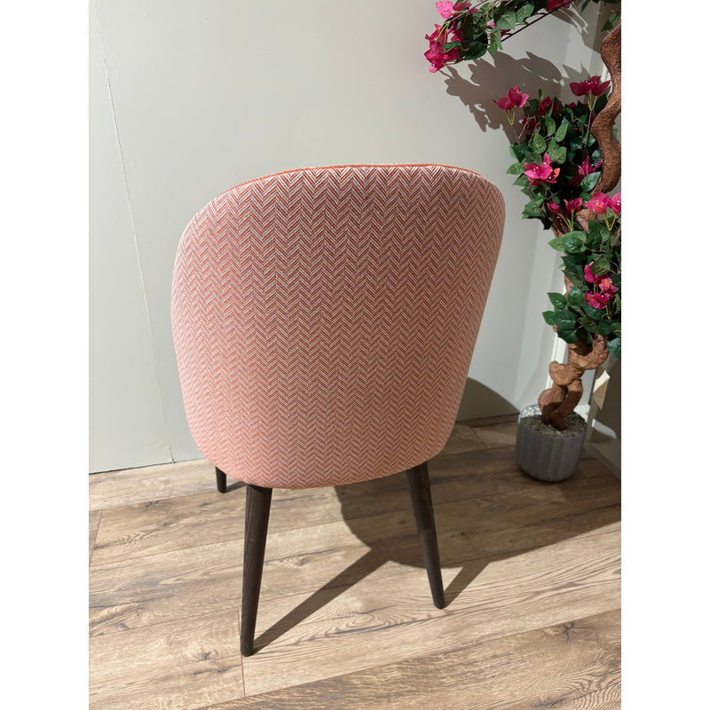 A Slim dining chair with pink herringbone upholstery and dark wooden legs stands on a wooden floor beside a green and pink flowering plant in a gray pot, facing away from the camera.