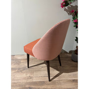 The Slim dining chair features a rust-orange seat, a light pink herringbone backrest, and dark wooden legs. This modern custom chair stands on a wooden floor next to a plant with pink flowers.