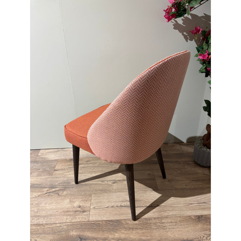 The Slim dining chair features a rust-orange seat, a light pink herringbone backrest, and dark wooden legs. This modern custom chair stands on a wooden floor next to a plant with pink flowers.