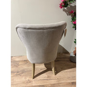 The Jacob chair, featuring light gray aqua clean fabric upholstery and wooden legs, is viewed from the back on a wooden floor next to a potted plant with pink flowers against a plain light-colored wall.