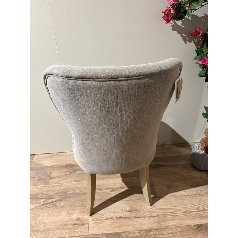 The Jacob chair, featuring light gray aqua clean fabric upholstery and wooden legs, is viewed from the back on a wooden floor next to a potted plant with pink flowers against a plain light-colored wall.