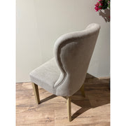 The Jacob chair, featuring a light gray curved backrest and grey washed wood legs, is placed on a wooden floor with its back to the camera. A plant with pink flowers appears partially in the upper right corner.