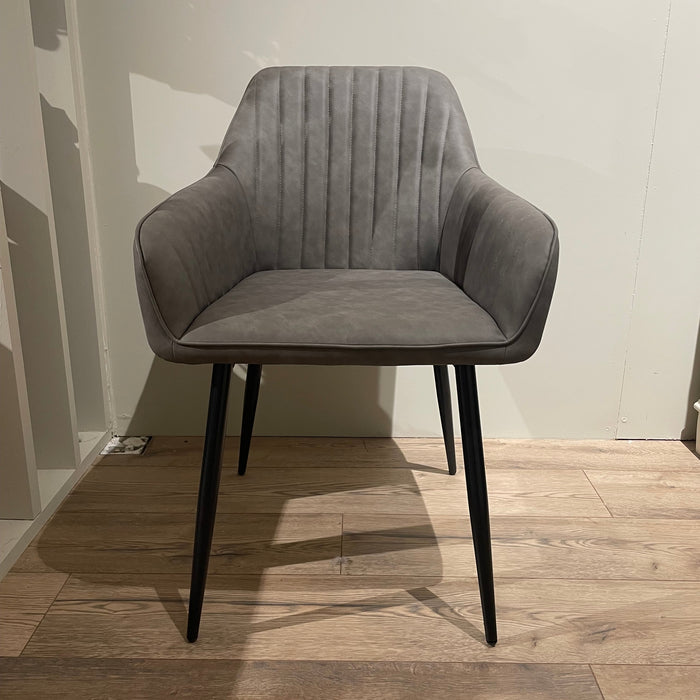 The Enzo Dining Chair features a modern gray design with leatherette cushioned seats, vertical stitching on the backrest, and black metal legs, elegantly placed on a wooden floor against a light-colored wall.