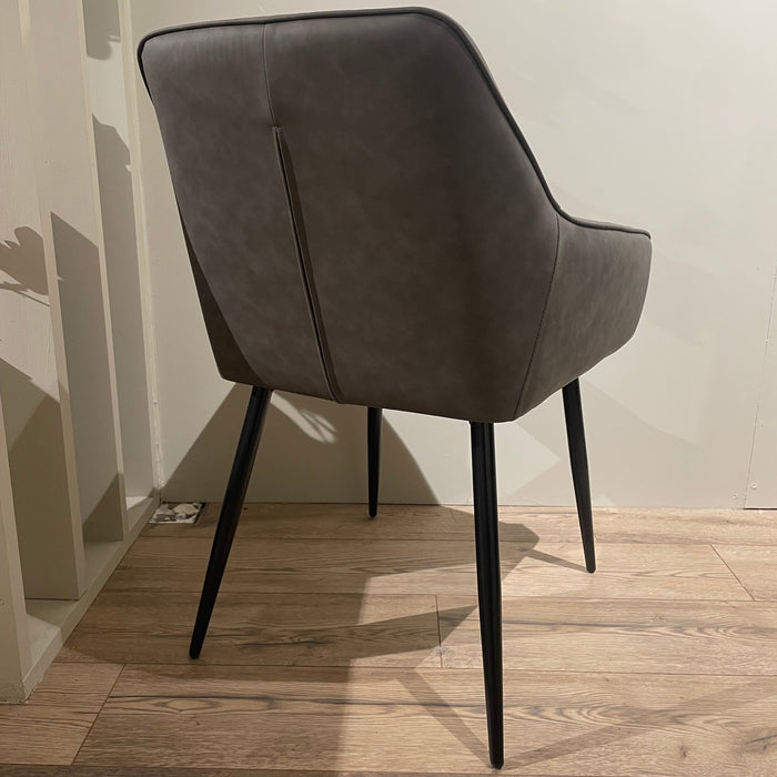 The Enzo Dining Chair features cushioned seats, a gray leatherette finish, a curved backrest, and slender black legs, displayed on a light wood floor against a plain, light-colored wall.