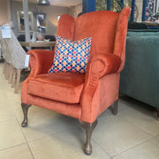 A Queen Anne Chair in burnt orange with dark wooden legs sits on tiled flooring, featuring a vibrant geometric cushion, displayed in a well-lit showroom.