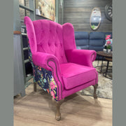 A vibrant pink Queen Anne Chair with floral patchwork fabric and wooden legs stands on a wood floor in a cozy, modern living room.