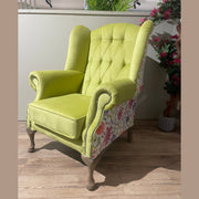 A lime green Queen Anne Chair with tufted back, winged sides, and patchwork outer fabric stands on wooden legs atop a light wood floor, accented by plants in the background—an elegant Irish-made piece.