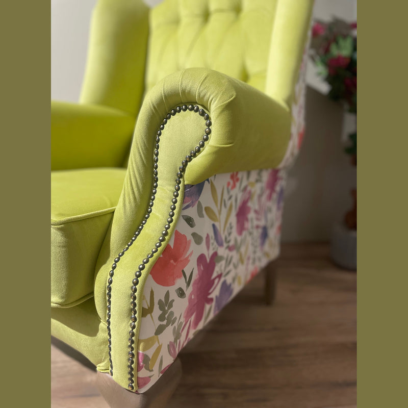 A close-up of the Queen Anne Chair features lime green upholstery, floral patterned sides, and decorative nailhead trim, set on a wooden floor with a blurred plant in the background.