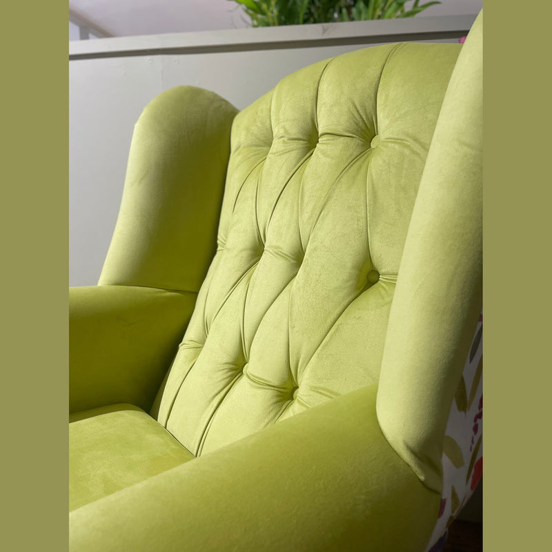 A close-up of the Queen Anne Chair, featuring light green tufted velvet upholstery, high sides, and a cushioned seat, shown indoors next to a partition with green plants in the background.