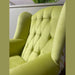 A close-up of the Queen Anne Chair, featuring light green tufted velvet upholstery, high sides, and a cushioned seat, shown indoors next to a partition with green plants in the background.