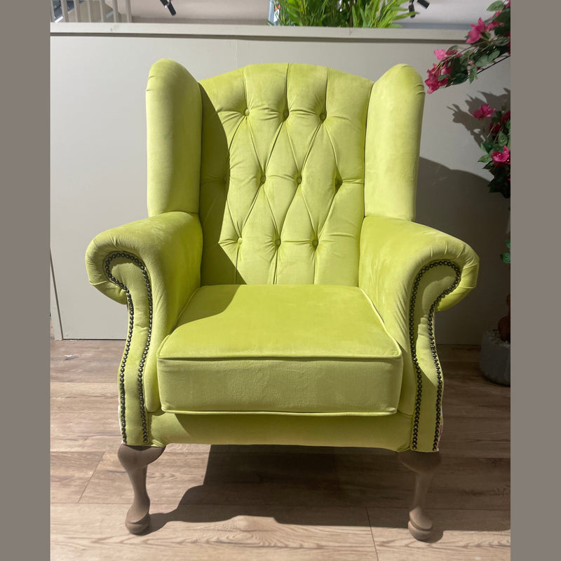 The Queen Anne Chair, in vibrant lime green with tufted upholstery, high back, rolled arms, nailhead trim, and wooden legs, sits on a light wood floor beside plants and flowers.