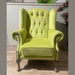 The Queen Anne Chair, in vibrant lime green with tufted upholstery, high back, rolled arms, nailhead trim, and wooden legs, sits on a light wood floor beside plants and flowers.