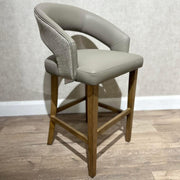 The Willow Counter Stool features a light gray upholstered seat and backrest, wooden legs, and a fabric accent on the back, set on a wooden floor against a textured light wall.