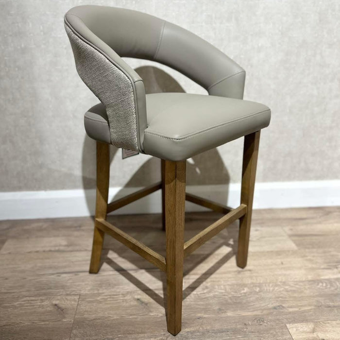 The Willow Counter Stool features a light gray upholstered seat and backrest, wooden legs, and a fabric accent on the back, set on a wooden floor against a textured light wall.