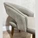 A close-up of the Willow Dining Chair showcases its light gray upholstery, textured outer backrest, and wooden legs—an elegant modern chair that stands out against a neutral wall and light wood floor.