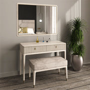 The Diletta Dressing Table has a marble top, ribbed drawers, an upholstered bench, large mirror, and decor accents next to a tall potted plant on a wooden floor in a bright bedroom.