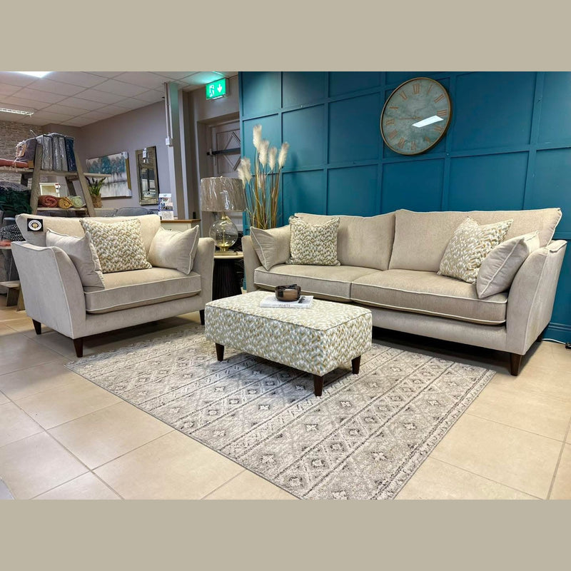 A modern living room showcases the Layla grand sofa and loveseat in washable aquaclean fabrics, accented with patterned pillows, a matching ottoman, decorative rug, large wall clock, and vase of tall dried grasses against a teal paneled wall.