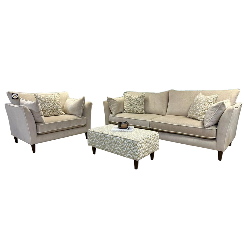 The Layla is a grand beige upholstered sofa set, Irish-made with wooden legs, a matching armchair, patterned pillows, and a rectangular washable aquaclean ottoman—all arranged on a white background.