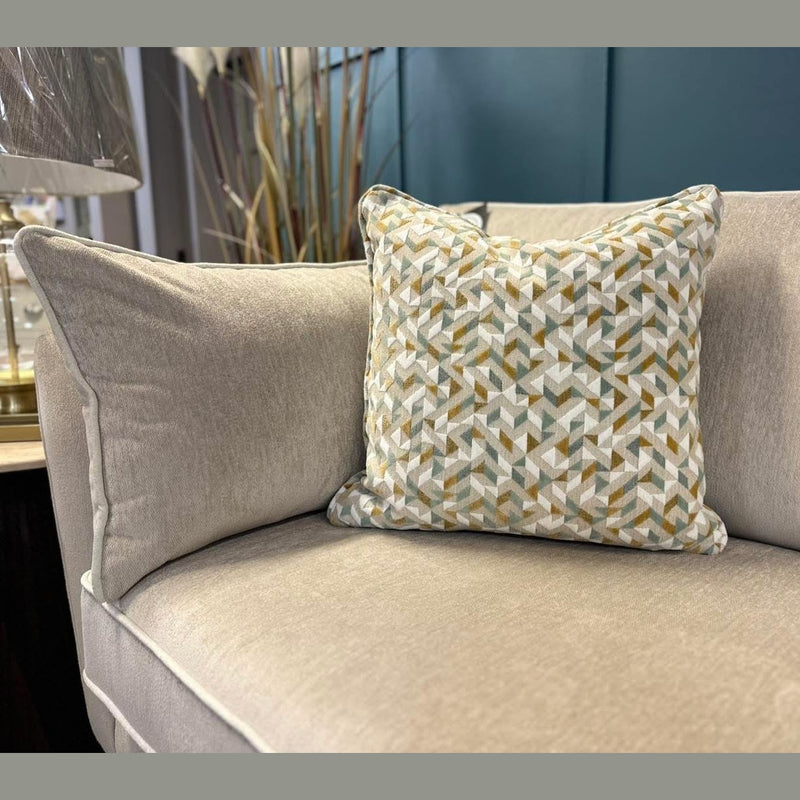 The Layla grand sofa, in beige and made in Ireland with washable aquaclean fabric, showcases a cushion with geometric patterns in muted green, gold, and white. Modern decor is seen in the background.