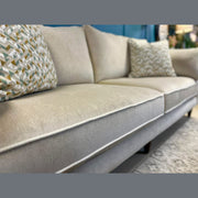 A close-up of the Layla sofa in light beige, made in Ireland with washable Aquaclean fabric. Two geometric-patterned neutral throw pillows sit on top, paired with a patterned rug and blue wall decor in the background.