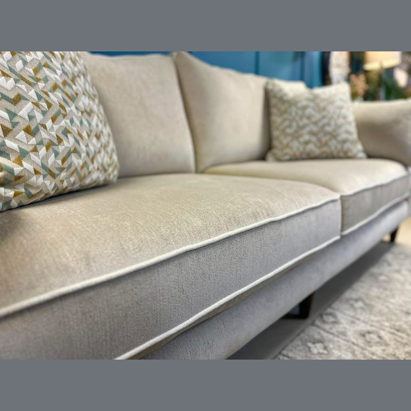 A close-up of the Layla sofa in light beige, made in Ireland with washable Aquaclean fabric. Two geometric-patterned neutral throw pillows sit on top, paired with a patterned rug and blue wall decor in the background.