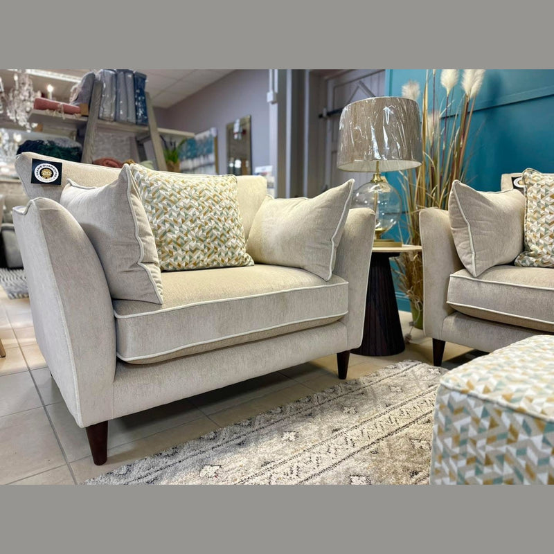 The Layla armchair in modern beige, featuring patterned cushions and Irish-made washable aquaclean fabric, sits on a light rug near its matching ottoman, pairing perfectly with the grand sofa and a glass lamp on the side table.