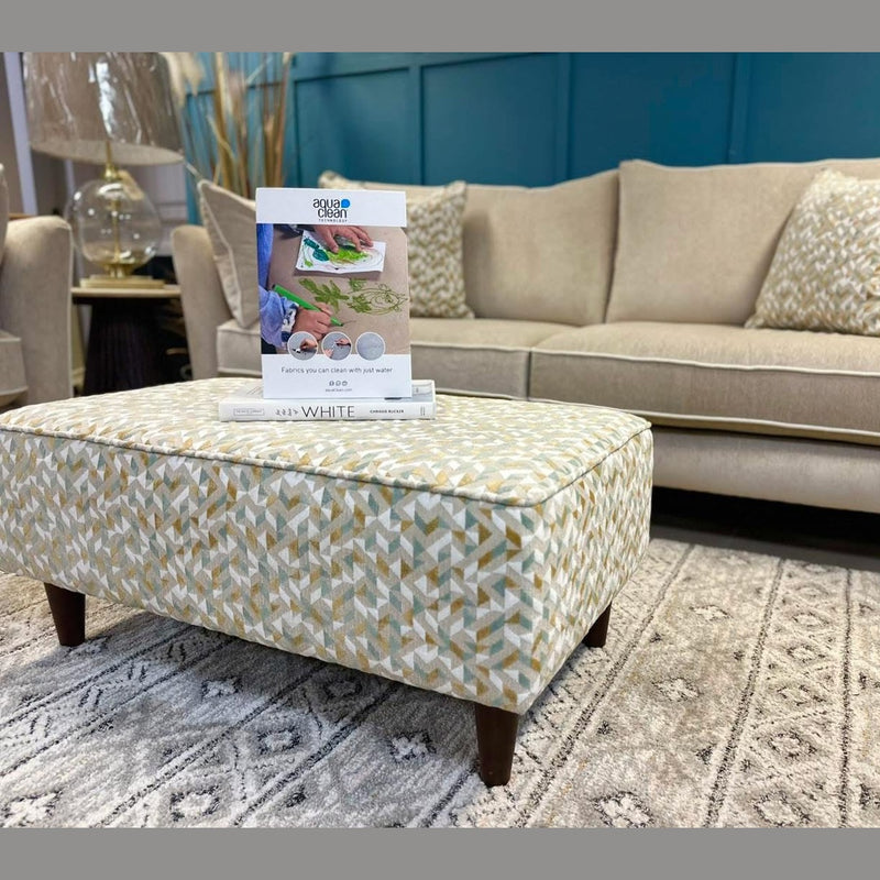 The Layla grand sofa with patterned pillows pairs with a matching ottoman, both in washable Aquaclean fabric, on a gray rug in a modern living room. A book or brochure is placed atop the ottoman.